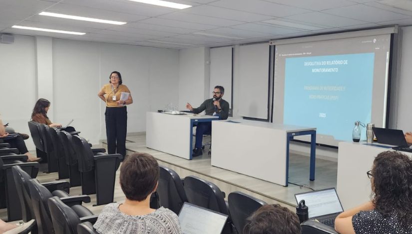 Fotografia do palco com um telão que mostra a imagem da capa de uma apresentação. Uma mulher em pé olhando para as pessoas da plateia e um homem sentado à mesa falando ao microfone.