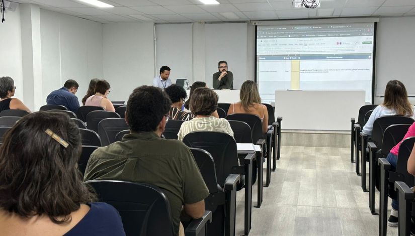 Fotografia mostra auditório com pessoas sentadas nas cadeiras e dois homens a frente do palco sentados à mesa com uma projeção em tela ao lado deles.