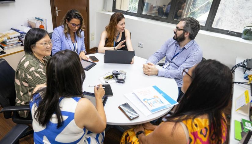 Fotografia de seis pessoas adultas sentadas ao redor da mesa conversando. Sobre a mesa papéis, computado e tablets.
