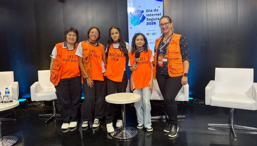  Três adolescentes e duas adultas usam colete laranja do programa "Imprensa Jovem" e olham para câmera posando para a foto.