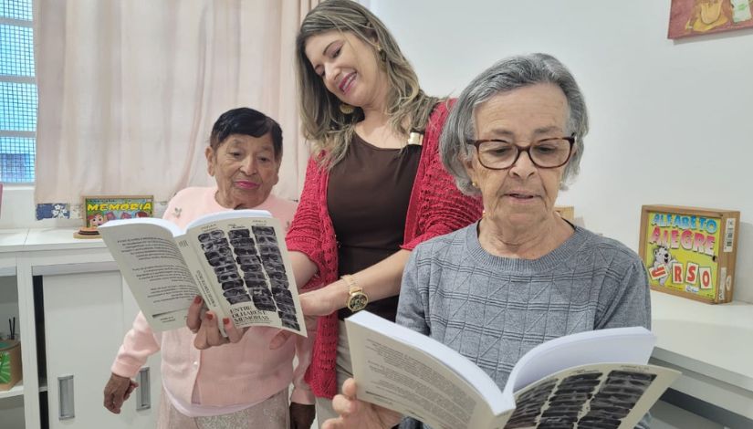 Fotografia de duas senhoras idosas segurando livros e uma mulher entre eles olhando o livro.
