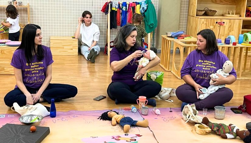 Fotografia de três mulheres sentadas no chão. A mulher que está ao centro segura uma boneca de pano e oferece leite em um copinho para a boneca durante uma oficina sobre extração de leite materno.