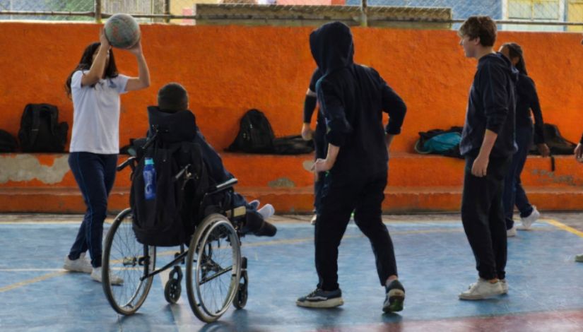 Estudantes participam de uma atividade de vôlei inclusivo em uma quadra escolar. Entre eles, um aluno em cadeira de rodas integra a dinâmica do jogo junto aos colegas, que acompanham o movimento da bola