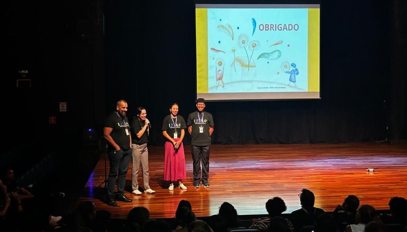Imagem mostra quatro pessoas em pé sobre um palco de madeira, em um auditório, durante um evento. Uma delas segura um microfone e parece estar falando ao público, enquanto as demais estão ao lado, sorrindo. Todas vestem camisetas escuras com identificação do evento. Ao fundo, há uma tela de projeção exibindo a palavra “OBRIGADO” acompanhada de ilustrações coloridas. Na parte inferior da imagem, é possível ver parte da plateia sentada, assistindo à apresentação. O ambiente é escuro, com iluminação direcionada ao palco, sugerindo o encerramento ou agradecimento de uma atividade.