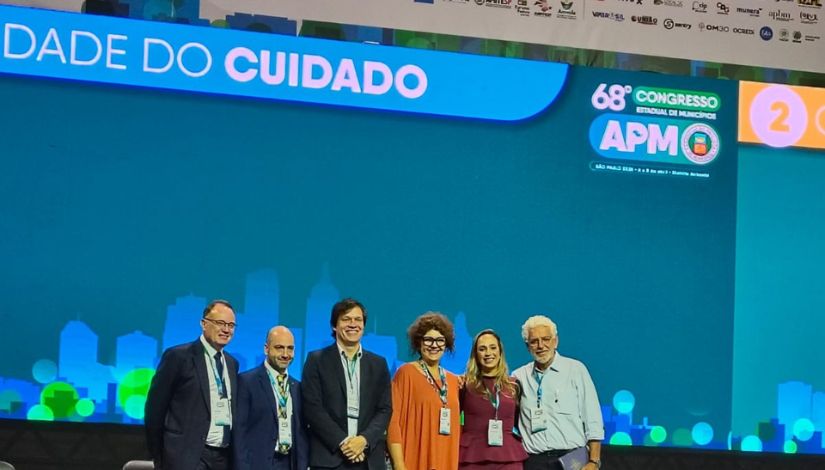 Imagem de um palco durante um evento institucional. Ao fundo, há um painel grande em tons de azul e verde a identificação do 68º Congresso Estadual de Municípios da APM. À frente do painel, seis pessoas estão lado a lado, em pé, sorrindo para a foto. Todas usam crachá de identificação. O grupo é composto por homens e mulheres, vestidos com trajes formais e sociais, como ternos, blazer e vestidos. O ambiente é bem iluminado, com destaque para o palco e os participantes.