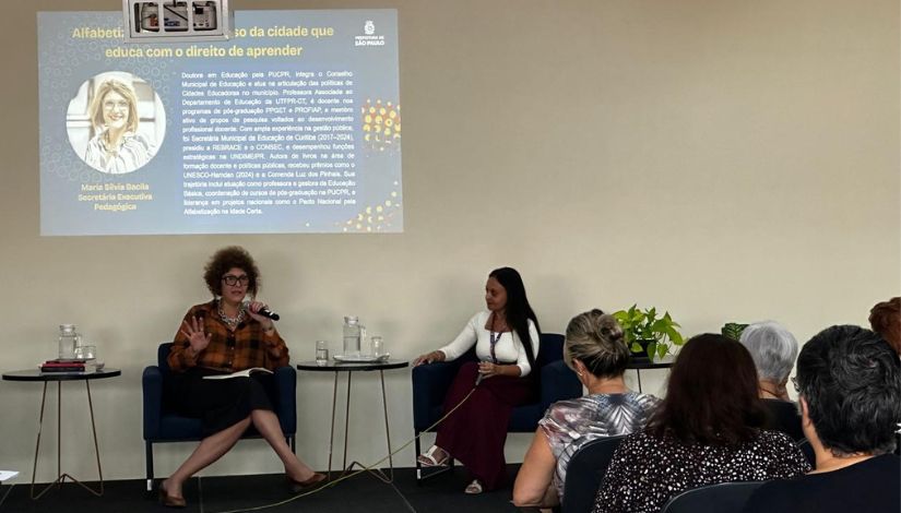 Imagem de um evento em ambiente interno, com duas mulheres sentadas em poltronas à frente de uma plateia. Uma delas fala ao microfone enquanto gesticula, e a outra escuta atentamente. Ao fundo, há uma projeção em tela com informações sobre alfabetização, incluindo texto institucional e a foto de uma mulher. Na frente das palestrantes, há pequenas mesas com copos e jarros de água. O público está sentado, de costas para a câmera, acompanhando a apresentação.