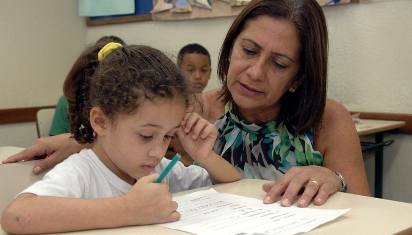 Imagem de uma criança pequena sentada à mesa, concentrada enquanto escreve ou desenha em uma folha de papel. Ao lado, uma mulher observa atentamente e oferece apoio, com expressão de cuidado e orientação. Ao fundo, outras pessoas aparecem desfocadas, sugerindo um ambiente escolar. A cena transmite um momento de aprendizagem e acompanhamento individual.