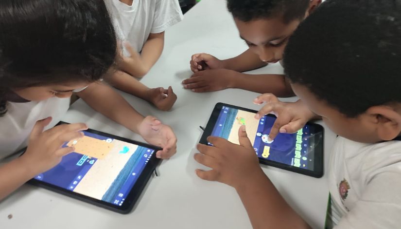 Imagem mostra quatro crianças reunidas em torno de uma mesa branca, utilizando tablets. Elas estão inclinadas para frente, concentradas nas telas, interagindo com um aplicativo colorido que apresenta elementos gráficos e personagens. As crianças vestem camisetas claras e utilizam as mãos para tocar e arrastar itens nos dispositivos. O ambiente é simples e bem iluminado, sugerindo um contexto escolar ou de atividade educativa com uso de tecnologia.