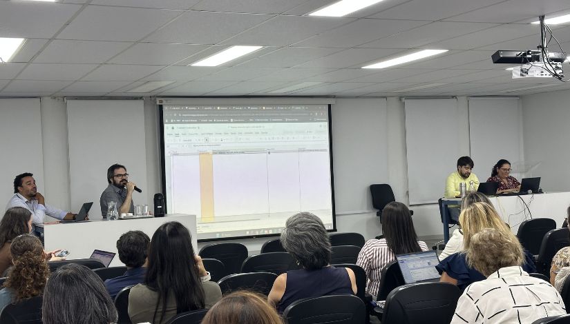 Fotografia de imagem projetada no telão. A frente um homem sentado e com microfone. Ele olha o telão e fala sobre a projeção.