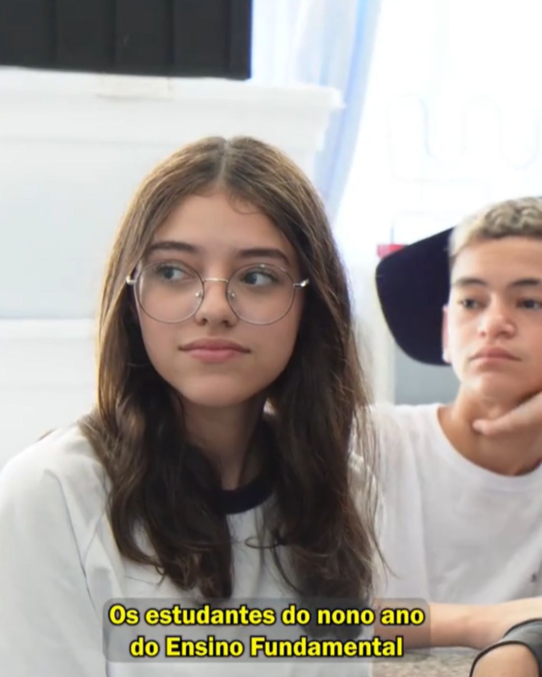 Imagem mostra dois adolescentes em uma sala de aula. Em primeiro plano, uma menina de cabelos castanhos, lisos e na altura dos ombros, usa óculos de grau com armação fina e camiseta clara. Ela está levemente virada para o lado, com expressão neutra e olhar atento. Ao fundo, um menino com cabelo curto aparece parcialmente, apoiando o queixo na mão, também com expressão séria. O ambiente é iluminado por luz natural, com paredes claras e uma janela ao fundo.