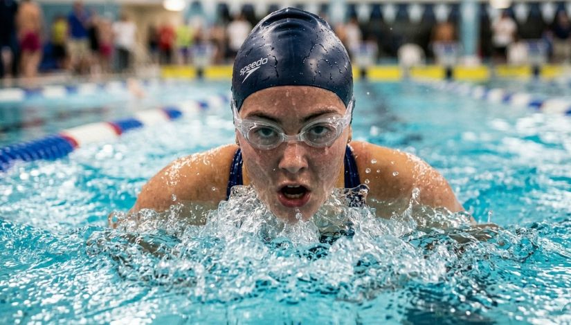 Imagem mostra uma nadadora em uma piscina durante a prática esportiva. Em primeiro plano, a atleta está parcialmente submersa, realizando um movimento de nado, com a cabeça fora d’água. Ela usa touca e óculos de natação e apresenta expressão de esforço e concentração, com a boca aberta enquanto respira. A água ao redor está agitada, com respingos visíveis. Ao fundo, é possível ver outras pessoas e raias da piscina, em ambiente que sugere treino ou competição em espaço aquático coberto.