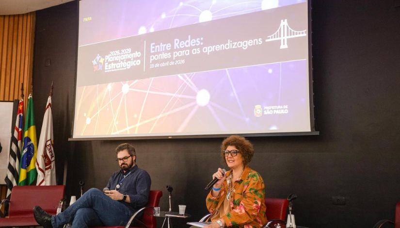 A imagem apresenta uma palestra ou painel de discussão ocorrendo em um auditório. Dois palestrantes estão sentados em poltronas vermelhas de couro à frente de um palco. À direita, uma mulher de cabelos castanhos curtos e cacheados, usando óculos de armação escura e uma jaqueta laranja com estampa floral colorida, fala ao microfone. À esquerda, um homem com barba e óculos, vestindo uma camisa azul escura e calça jeans, observa a colega enquanto segura um objeto. Uma grande tela de projeção exibe um slide com fundo roxo e azul, decorado com linhas conectadas que lembram uma rede neural ou digital. No centro da tela, lê-se o título: 