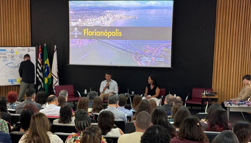 A imagem captura um auditório durante uma palestra ou evento institucional. O ambiente é formal e bem estruturado, com foco em um palco onde dois palestrantes estão sentados. Um homem e uma mulher estão sentados em poltronas vermelhas de estilo moderno. O homem, à esquerda, veste uma camisa branca e segura um microfone, parecendo estar falando. A mulher, à direita, veste uma blusa preta e observa o colega. Vê-se a parte de trás das cabeças e ombros de uma plateia diversificada, composta por várias pessoas sentadas em poltronas escuras. O público está atento e voltado para a frente.