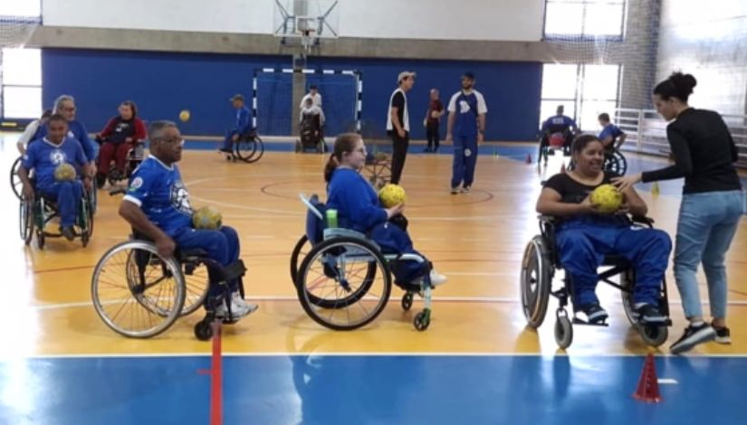 Fotografia de pessoas em cadeira de rodas praticantes de handbol adaptado. Eles estão se deslocando em um grande círculo com a bola no colo.