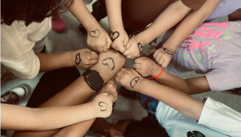 Vista superior de várias crianças reunidas em círculo, com os punhos encostados no centro. Em cada mão há um desenho simples de coração feito com caneta, simbolizando união e afeto. As crianças usam roupas casuais e estão próximas umas das outras, formando uma composição que transmite coletividade, amizade e colaboração.