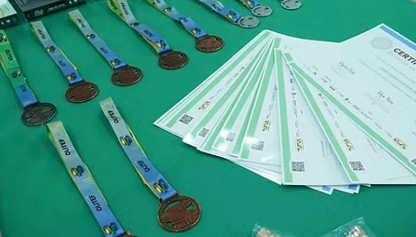Imagem mostra uma mesa com fundo verde onde estão dispostas medalhas e certificados. À esquerda, várias medalhas com fitas azuis estão organizadas em fileiras. À direita, há um conjunto de certificados empilhados em formato de leque, com detalhes em verde. A composição destaca materiais de premiação, sugerindo um evento de reconhecimento ou competição educacional.