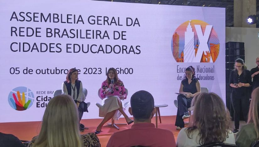 No palco, três mulheres estão sentadas em poltronas brancas modernas: A mulher ao centro fala ao microfone. Ela veste uma blusa rosa vibrante com estampas e uma saia branca. À esquerda, uma mulher observa a fala, vestindo uma blusa branca de mangas compridas sob um colete escuro. À direita, outra mulher segura papéis e parece estar acompanhando a mediação ou tomando notas, vestindo uma blusa preta sem mangas. A foto foi tirada da perspectiva da plateia, sendo possível ver o topo da cabeça de algumas pessoas em primeiro plano, o que confere uma sensação de profundidade e participação no evento.