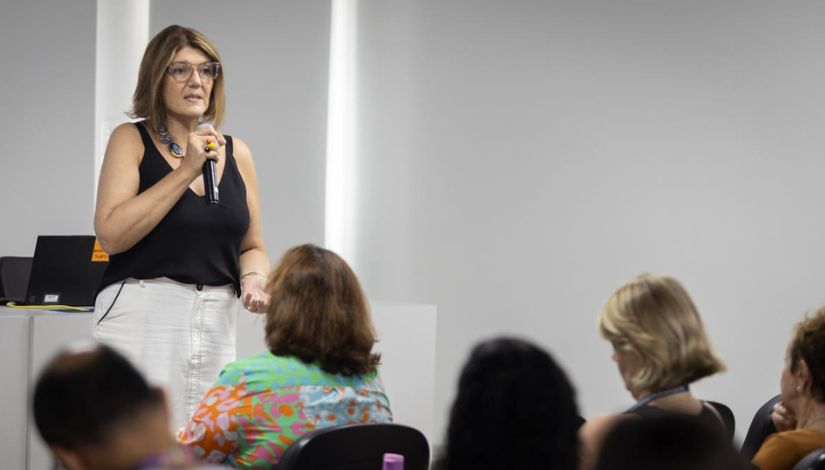 Imagem mostra uma mulher adulta em pé, em um ambiente interno, durante uma apresentação. Ela segura um microfone com uma das mãos e gesticula com a outra, demonstrando postura de fala e condução. Veste blusa preta sem mangas e calça clara, além de usar óculos. À sua frente, há pessoas sentadas, vistas de costas, que acompanham a apresentação com atenção. O ambiente é claro, com paredes neutras, sugerindo um espaço de formação, palestra ou seminário.