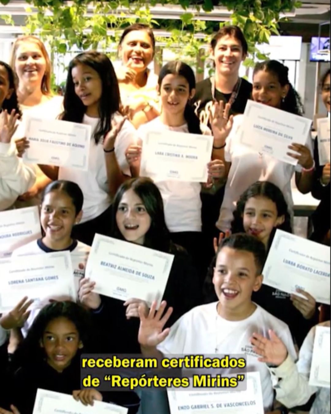 Imagem mostra um grupo de crianças e alguns adultos reunidos em um ambiente interno, sorrindo e segurando certificados. As crianças estão posicionadas em diferentes alturas, algumas em pé e outras mais à frente, todas com expressões de alegria e orgulho. Os certificados são exibidos para a câmera, indicando uma cerimônia de reconhecimento. Ao fundo, há plantas e iluminação natural, sugerindo um espaço acolhedor. Na parte inferior da imagem, aparece o texto: “receberam certificados de ‘Repórteres Mirins’”