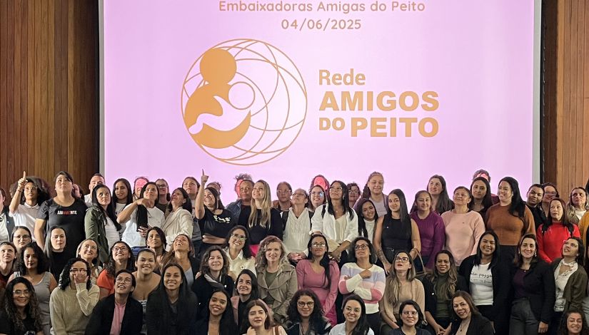 Fotografia de muitas mulheres posando para câmera. Ao fundo, projeto no telão o texto 