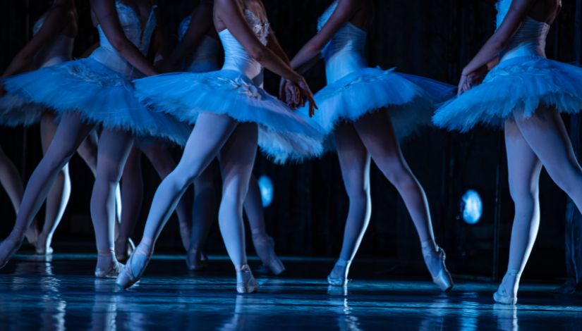 Fotografia de bailarinas dançando no palco. Elas aparecem dos ombros para baixo.