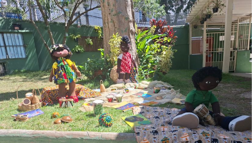 Fotografia de bonecos negros sobre panos cercados de livros e instrumentos musicas disposto da aárea externa da escola. Espaço possui grama verde e árvores.