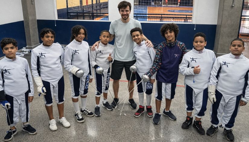 Fotografia de um professor ao centro e estudantes ao seu lado direito e esquerdo com roupa de esgrima.