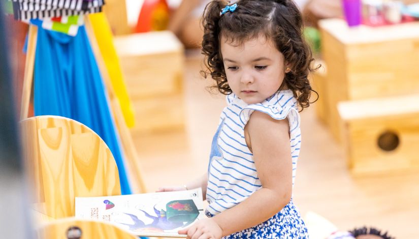 Fotografia de uma criança com um livro nas mãos. Ela olha para o lado e ao redor dela há brinquedos em madeira.