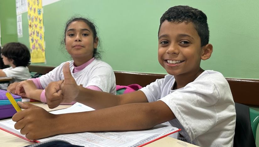 Duas crianças, uma menina e um menino, sentado na sala de aula. Eles olham para câmera e sorriem, o menino faz sinal de positivo com o polegar direito para cima.