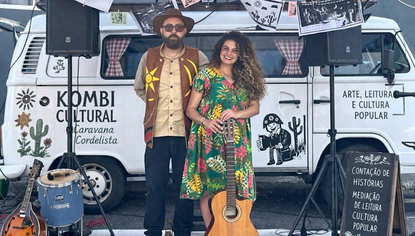 Fotografia de uma kombi com várias gravura e inscrições e um casal a frente vestidos para apresentação cultura com chapéu, vestido e violão.