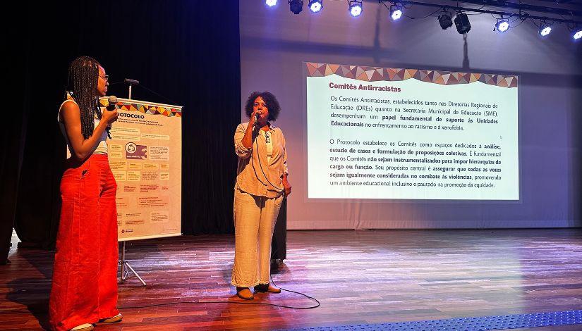 Fotografia de duas mulheres negras no palco. Ao fundo uma projeção com informações sobre os Comitês Antirracistas.