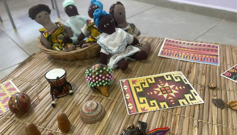 Sobre uma esteira de palha, estão dispostos elementos que remetem às culturas afro-brasileira e indígena, como bonecas de pano negras com roupas coloridas, instrumentos musicais, sementes, objetos artesanais e cartões com estampas geométricas em tons terrosos. Ao fundo, algumas bonecas estão acomodadas em um cesto de vime, compondo um ambiente educativo e cultural.