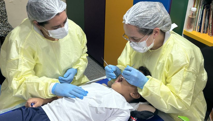 Fotografia de duas dentistas com o avental amarelo, máscara e toca branca. Elas estão atendendo uma criança que está deitada na maca.