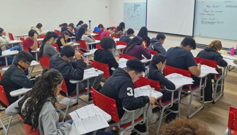 Sala de aula com estudantes sentados em cadeiras realizando uma atividade escrita. Os estudantes estão concentrados, lendo e preenchendo folhas de exercícios. À frente da sala, há um quadro branco com anotações feitas pela professora, compondo um ambiente de estudo e preparação acadêmica.
