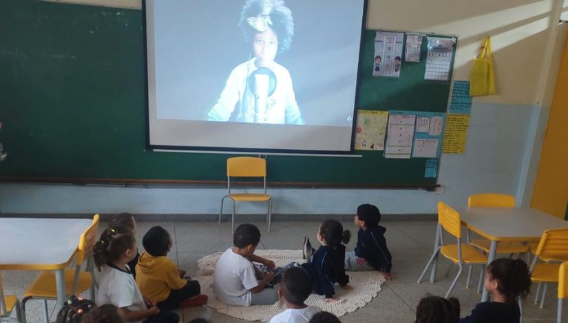 Crianças pequenas sentadas no chão de uma sala de aula assistem atentamente a uma projeção em tela. A imagem projetada mostra uma criança negra em destaque. O ambiente possui cadeiras e mesas amarelas, lousa verde ao fundo e cartazes pedagógicos fixados na parede, compondo um espaço escolar acolhedor e educativo.
