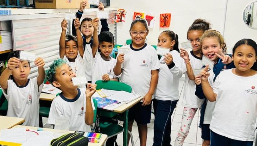 Fotografia de várias crianças com uniforme escolar da PMSP. Elas estão na sala de aula e seguram carteirinhas em suas mãos.