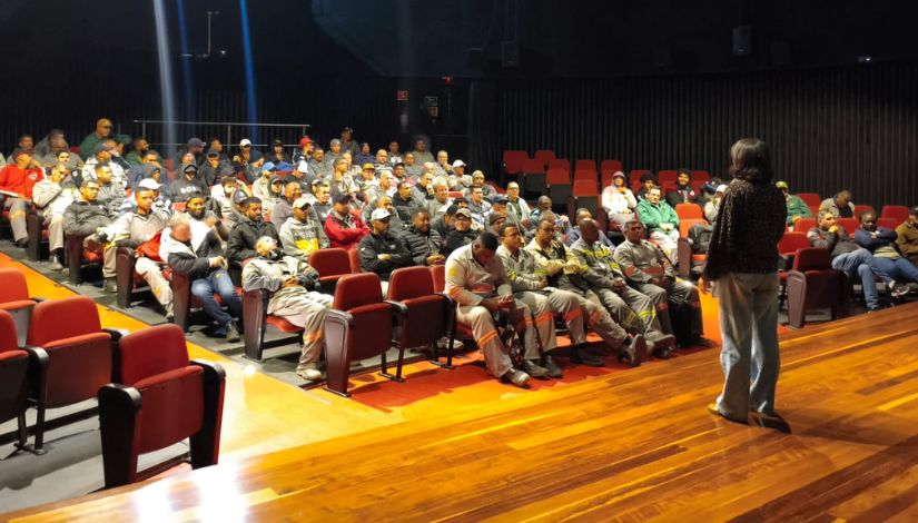Fotografia de um auditório durante uma formação profissional. Em primeiro plano, uma mulher está em pé sobre o palco de madeira, de costas para a câmera, conduzindo a atividade. À sua frente, dezenas de participantes ocupam as poltronas vermelhas do auditório, acompanhando atentamente a apresentação. A maioria dos presentes veste uniformes de trabalho nas cores cinza e laranja, enquanto outros utilizam roupas casuais. O ambiente possui iluminação direcionada para o palco e arquibancadas em diferentes níveis, criando um cenário de encontro formativo e troca de conhecimentos entre os profissionais.