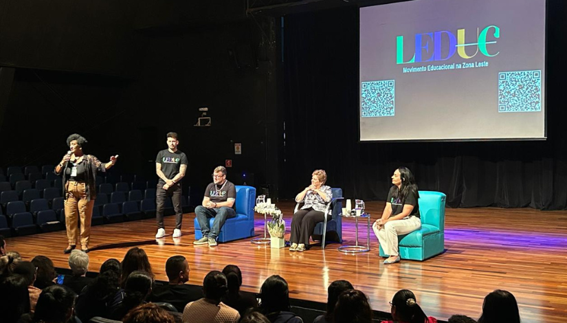 Auditório com palco de madeira durante um evento educacional do Movimento LEDUC. À esquerda, uma mulher negra fala ao microfone para o público. No centro do palco, um homem está em pé e duas mulheres e um homem estão sentados em poltronas azuis. Ao fundo, um telão exibe a logo colorida do LEDUC e QR Codes nas laterais. O público acompanha atentamente a atividade.