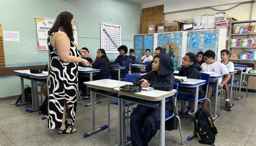 Fotografia de uma professora a frente dos seus alunos na sala de aula. Eles usam uniforme escolar da Prefeitura de São Paulo.