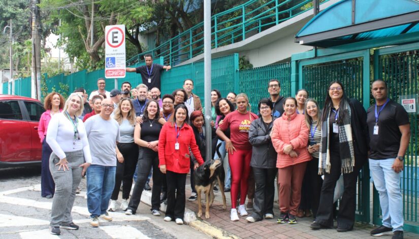 Um grupo de pessoas posa para uma foto em frente ao prédio da Diretoria Regional de Educação Guaianases. Os participantes estão reunidos lado a lado, sorrindo para a câmera. No centro do grupo, há um cachorro cercado pelas pessoas, tornando a cena mais acolhedora e descontraída. Ao fundo, é possível ver a fachada da instituição e a entrada do local.