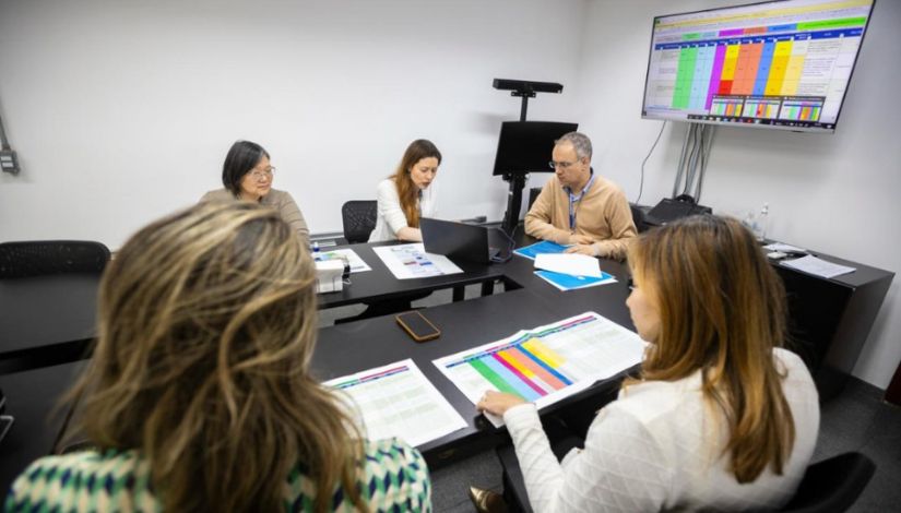 pessoas reunidas ao redor de uma mesa, em reunião. No projetor da parede e sobre a mesa, consta uma planilha com colunas coloridas