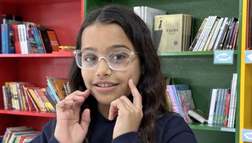 Menina de óculos. Ao fundo, prateleira com livros.