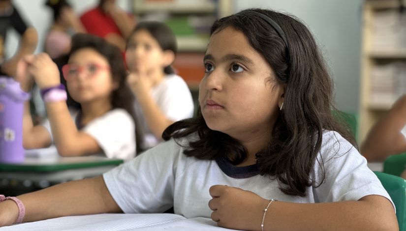 Fotografia de uma menina em primeiro plano. Ela está focada olhando para frente com o caderno sobre a mesa.