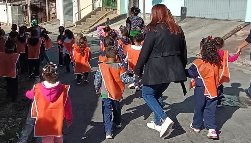 Fotografia de crianças usando coletes laranjas caminhando na rua com pessoas adultas.