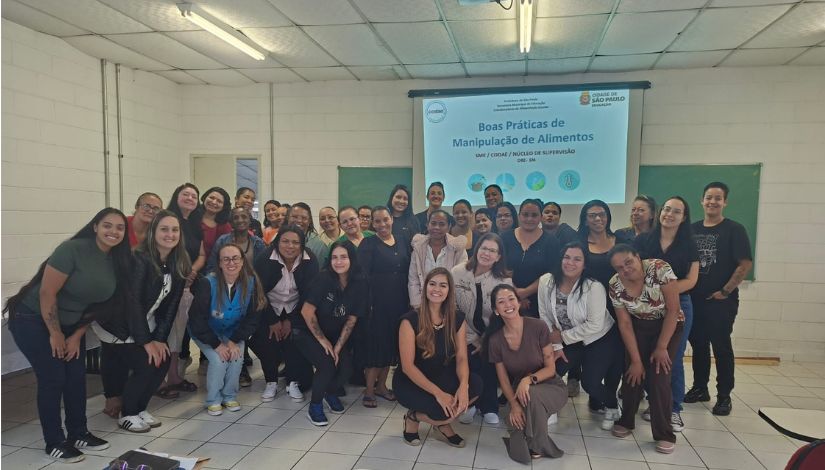 Fotografia em plano médio e ambiente fechado mostra um grupo de cerca de 30 mulheres reunidas em uma sala de aula ou treinamento. Elas estão posicionadas à frente de uma tela de projeção e sorriem para a câmera.