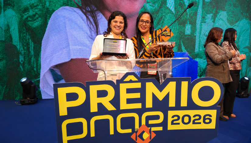Duas mulheres sorriem no palco durante uma cerimônia de premiação, posicionadas atrás de um púlpito transparente. Uma delas segura uma placa de homenagem e a outra exibe um troféu. À frente, há um grande letreiro com a inscrição 