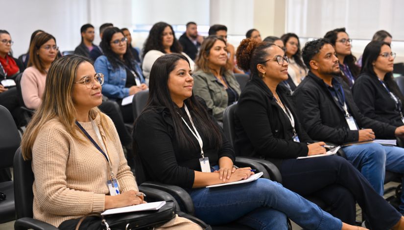 Grupo de adultos participa de um encontro de formação em uma sala de capacitação. Os participantes estão sentados em fileiras, atentos à apresentação, alguns com cadernos e canetas para anotações. O ambiente é amplo, iluminado e organizado, favorecendo o aprendizado e a integração dos novos profissionais.