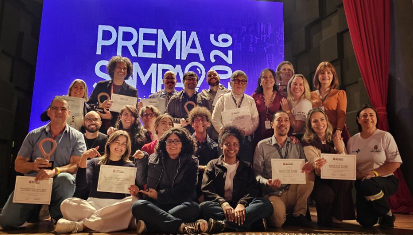 Fotografia com os vencedores do Premia Sampa 2026 no palco. Eles seguram certificados. Ao fundo, telão com a projeção da logomarca do 
