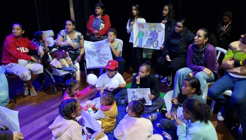 Fotografia de adultos e crianças sentados em um palco em momento de roda de conversa, alguns integrantes seguram cartazes.