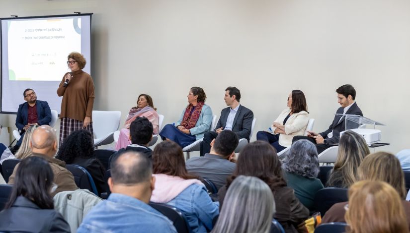 Fotografia de um encontro formativo realizado em auditório. Em primeiro plano, participantes sentados acompanham a atividade. Ao fundo, uma mulher em pé segura um microfone e fala ao público, enquanto outras seis pessoas estão sentadas em poltronas compondo uma mesa de debate. Ao lado do grupo, há uma tela de projeção com a identificação do evento. O ambiente é amplo, iluminado e reúne educadores e gestores em momento de formação e troca de experiências.